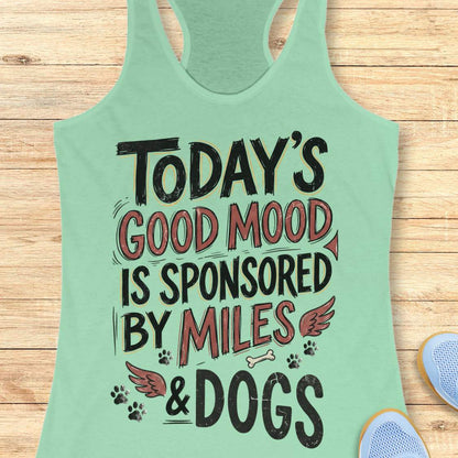 A mint green racerback tank top laid flat on a light wood background, featuring a distressed graphic that reads 'TODAY'S GOOD MOOD IS SPONSORED BY MILES & DOGS' with dog paw prints, wings, and a small bone. Part of a blue shoe is visible at the bottom right.