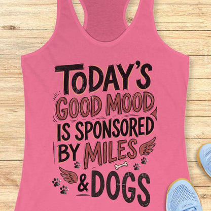 A pink racerback tank top laid flat on a light wood background, featuring the text 'TODAY'S GOOD MOOD IS SPONSORED BY MILES & DOGS' in black and brown lettering with small paw prints, wings, and a bone graphic. Partial blue shoes are visible at the edges.