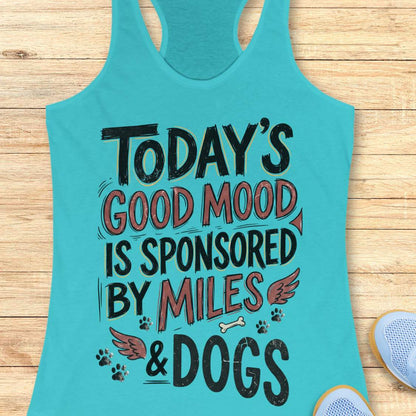 A turquoise racerback tank top laid flat on a light wood surface, featuring the printed phrase “TODAY'S GOOD MOOD IS SPONSORED BY MILES & DOGS” in black and muted red lettering with small dog-themed graphics. Part of a light blue shoe is visible at the lower right edge.