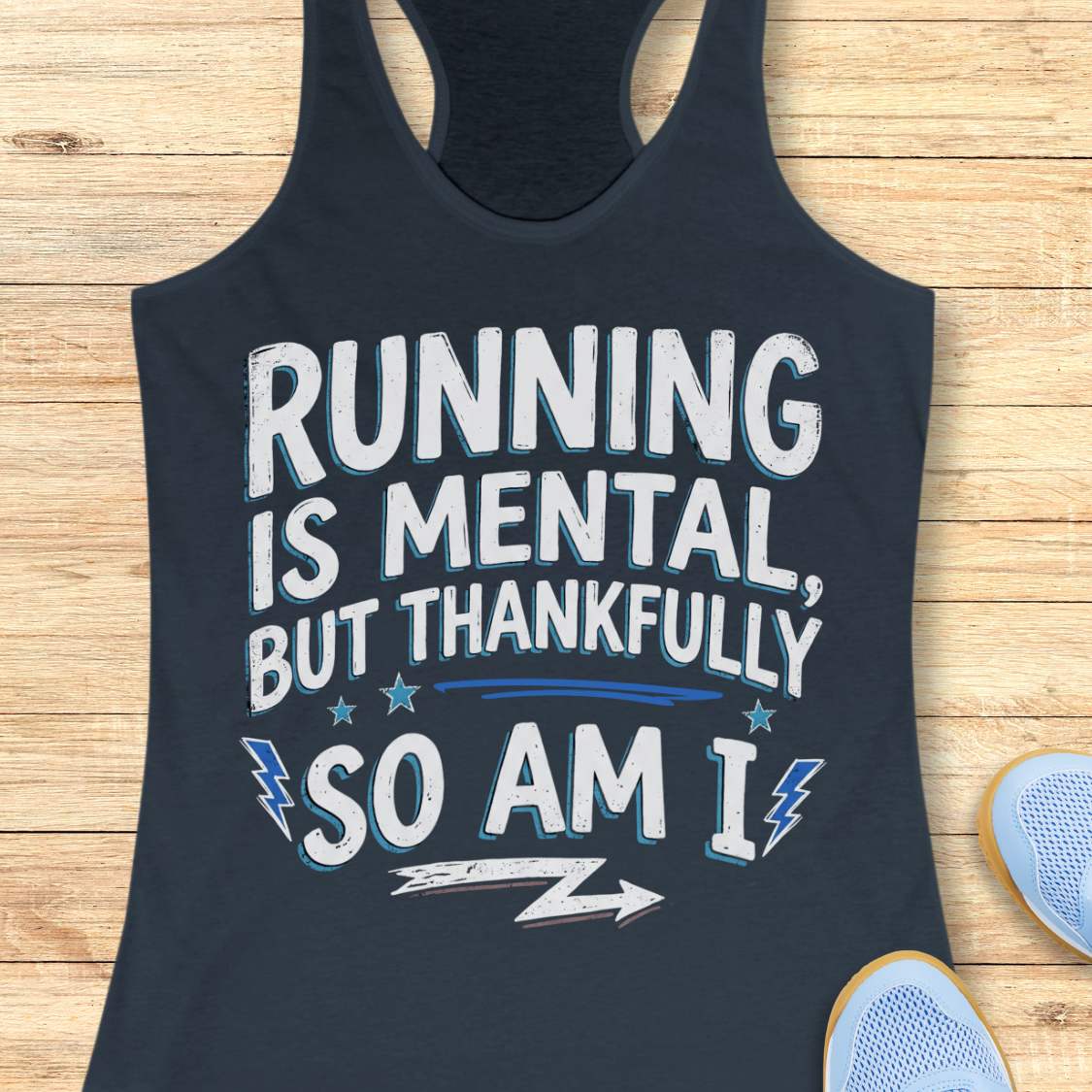 A indigo racerback tank top with a motivational graphic reading 'RUNNING IS MENTAL, BUT THANKFULLY SO AM I' in white and light blue, laid flat on a light wood plank background with partial sneakers visible at the edges.