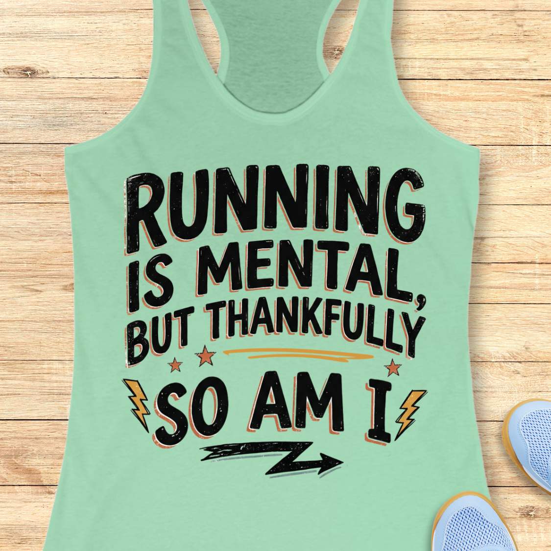 A mint green racerback tank top laid flat on a light wood background, featuring a motivational graphic that reads 'RUNNING IS MENTAL, BUT THANKFULLY SO AM I' in black with orange and yellow accents. Partial blue sneakers are visible at the bottom right as styling props.