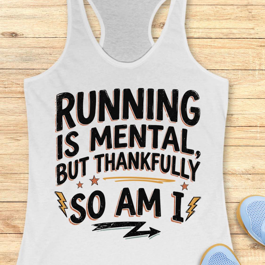 A white racerback tank top laid flat on a light wood plank background, featuring the printed phrase 'RUNNING IS MENTAL, BUT THANKFULLY SO AM I' in black lettering with small orange and yellow graphic accents. Partial blue sneakers are visible at the bottom right as styling props.