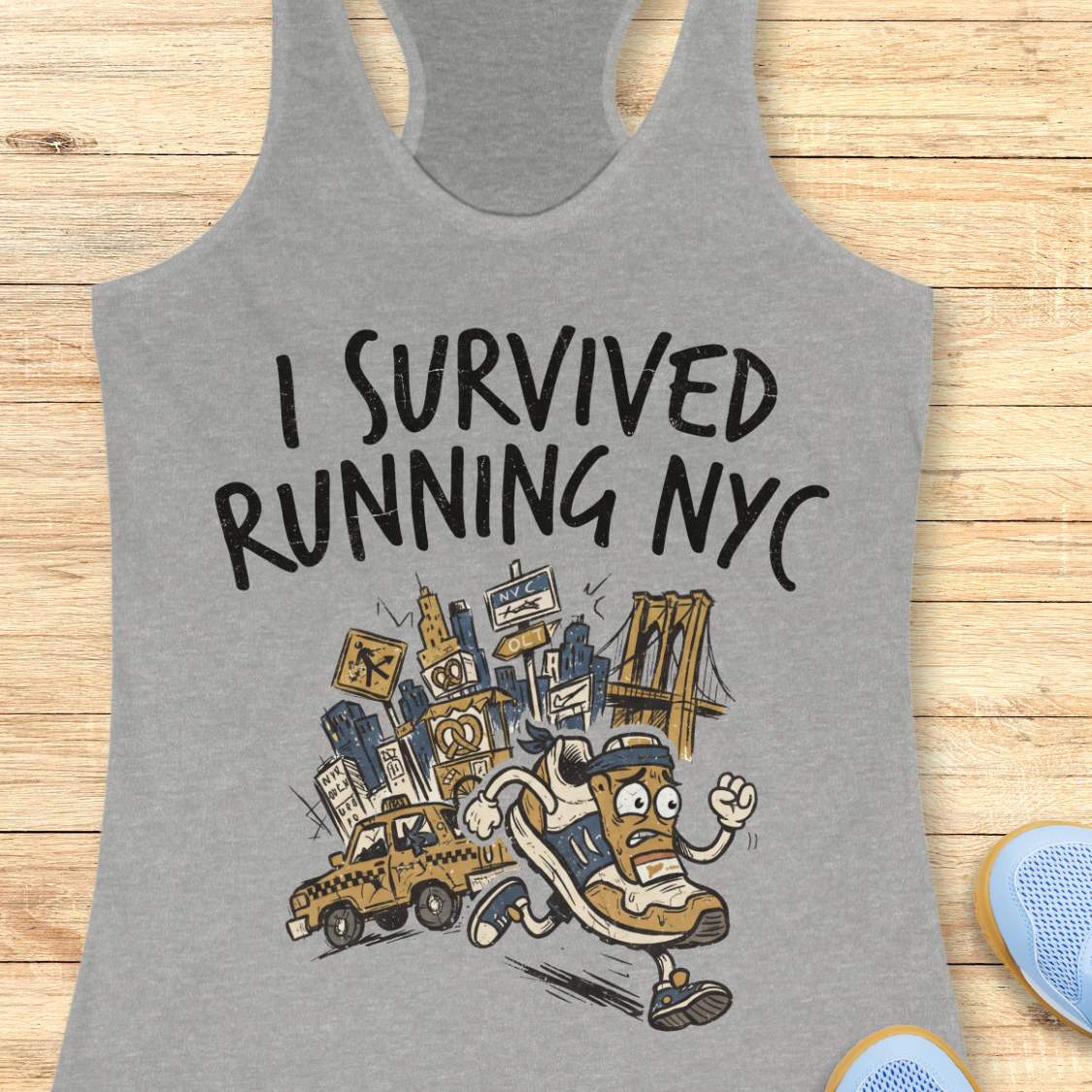 A heather gray racerback tank top laid flat on a light wood background, featuring a graphic print that reads 'I SURVIVED RUNNING NYC' above an illustrated cartoon figure running through city landmarks and a taxi.