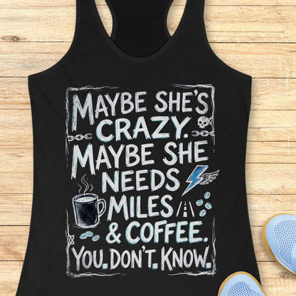A black racerback tank top laid flat on a light wood background, featuring a large white and blue graphic text that reads, 'MAYBE SHE'S CRAZY. MAYBE SHE NEEDS MILES & COFFEE.