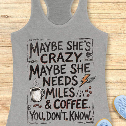 A grey racerback tank top laid flat on a light wood background, featuring a large white and blue graphic text that reads, 'MAYBE SHE'S CRAZY. MAYBE SHE NEEDS MILES & COFFEE.
