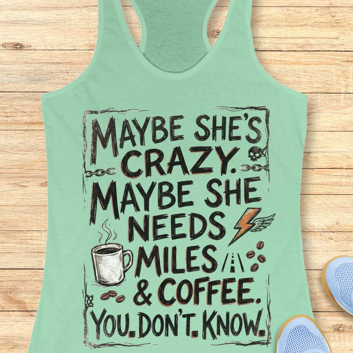 A mint green racerback tank top with a humorous graphic print, displayed flat on a light wood background with a pair of blue sneakers partially visible at the bottom right.
