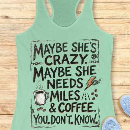 A mint green racerback tank top with a humorous graphic print, displayed flat on a light wood background with a pair of blue sneakers partially visible at the bottom right.