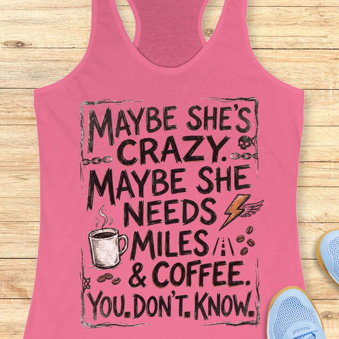 A pink racerback tank top laid flat on a light wood background, featuring a black graphic text design that reads 'MAYBE SHE'S CRAZY. MAYBE SHE NEEDS MILES & COFFEE.