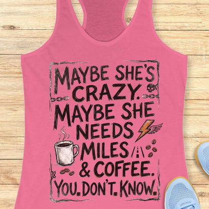 A pink racerback tank top laid flat on a light wood background, featuring a black graphic text design that reads 'MAYBE SHE'S CRAZY. MAYBE SHE NEEDS MILES & COFFEE.