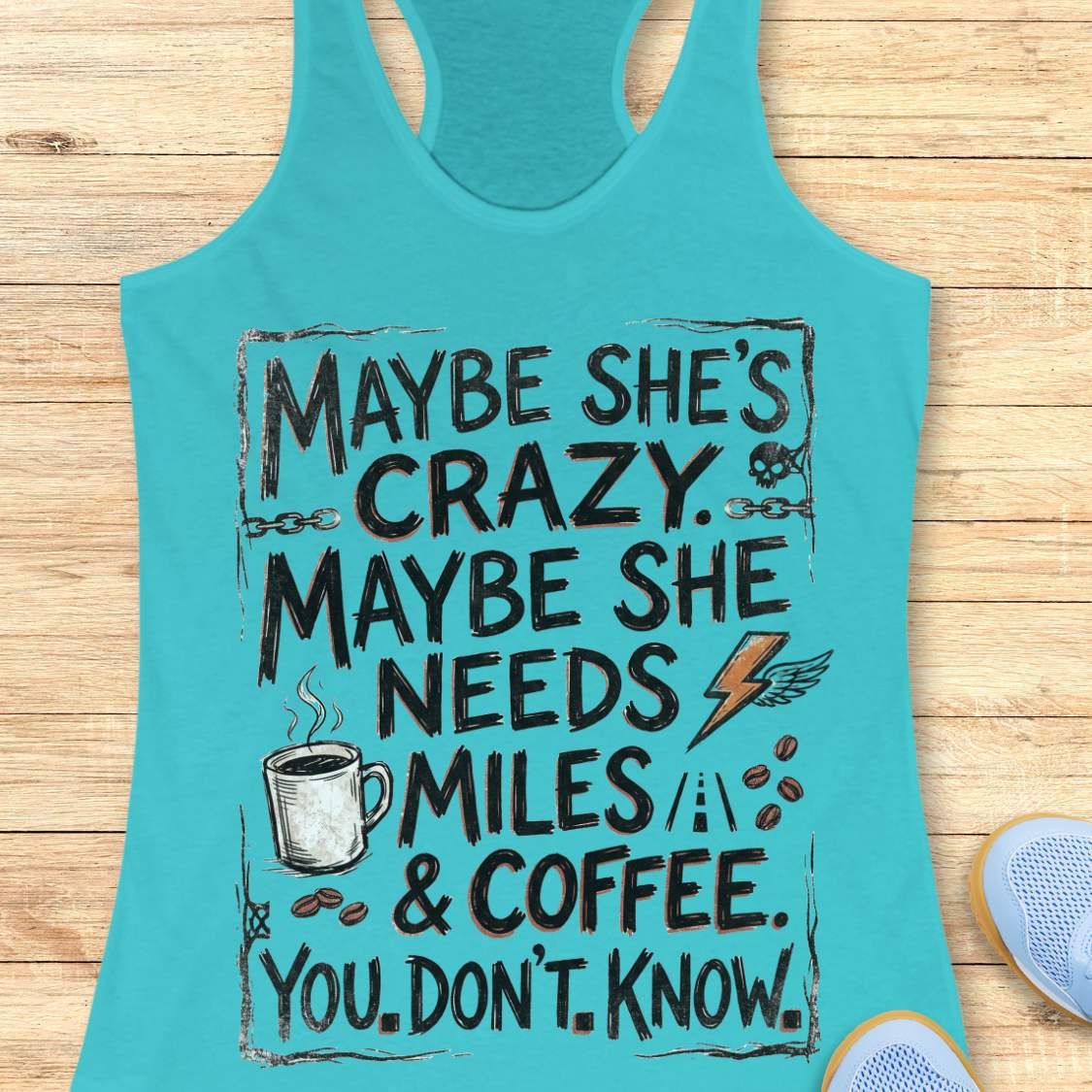 A blue racerback tank top laid flat on a light wood background, featuring a large white and blue graphic text that reads, 'MAYBE SHE'S CRAZY. MAYBE SHE NEEDS MILES & COFFEE.