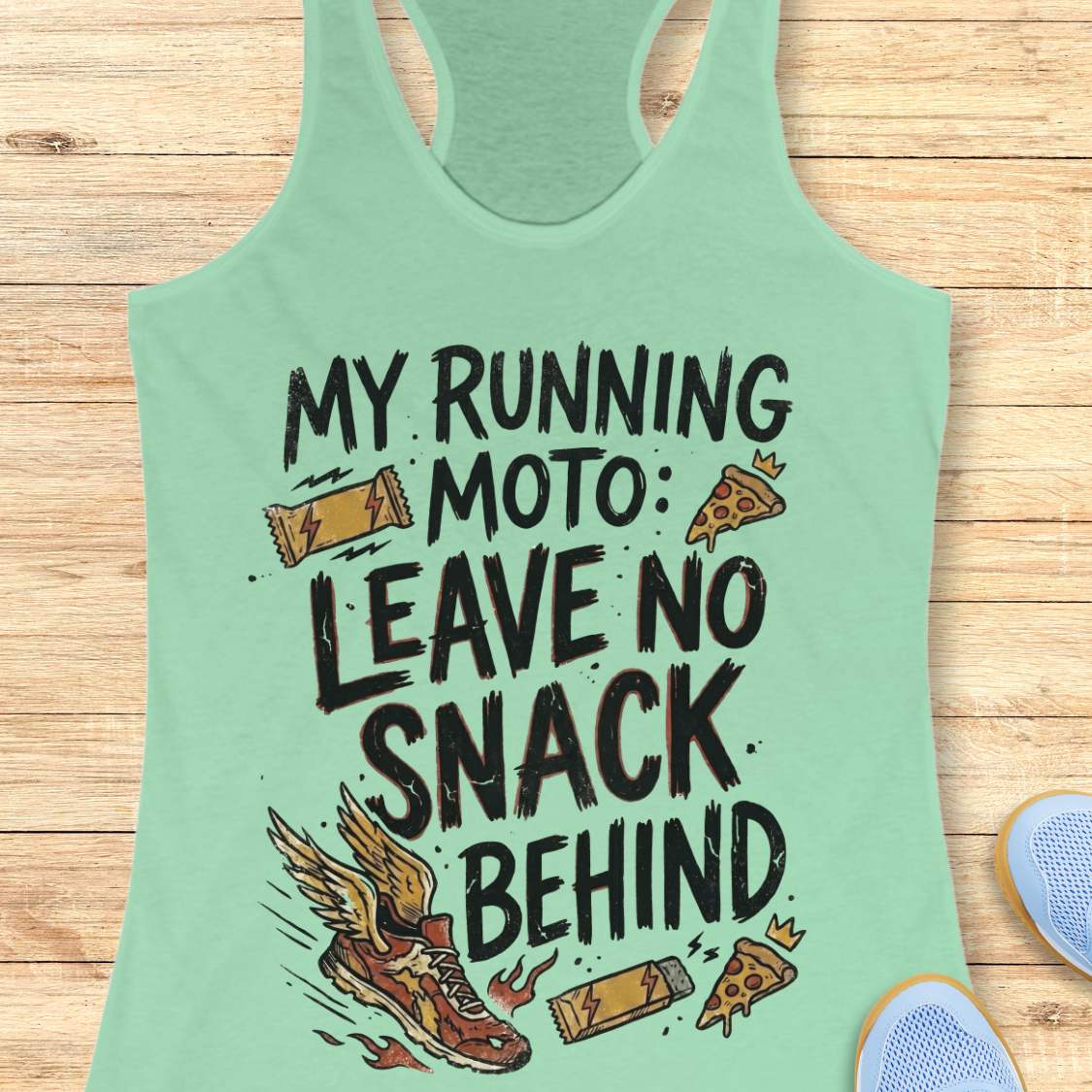 A mint green racerback tank top with a humorous graphic print reading 'MY RUNNING MOTO: LEAVE NO SNACK BEHIND' in black text, decorated with illustrations of a winged running shoe, a slice of pizza, and candy bars. The tank top is laid flat on a light brown wooden plank background, with partial blue sneakers visible in the bottom right corner.