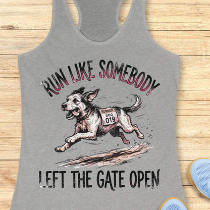 A heather gray racerback tank top laid flat on a light wood background, featuring a graphic of a dog wearing a race bib and the text 'RUN LIKE SOMEBODY LEFT THE GATE OPEN'.