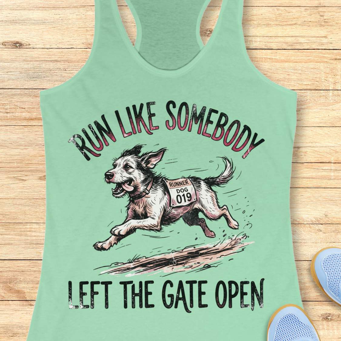A mint green racerback tank top laid flat on a light wood background, featuring a graphic of a running dog with the text 'RUN LIKE SOMEBODY LEFT THE GATE OPEN'.