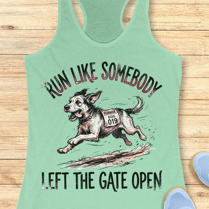 A mint green racerback tank top laid flat on a light wood background, featuring a graphic of a running dog with the text 'RUN LIKE SOMEBODY LEFT THE GATE OPEN'.