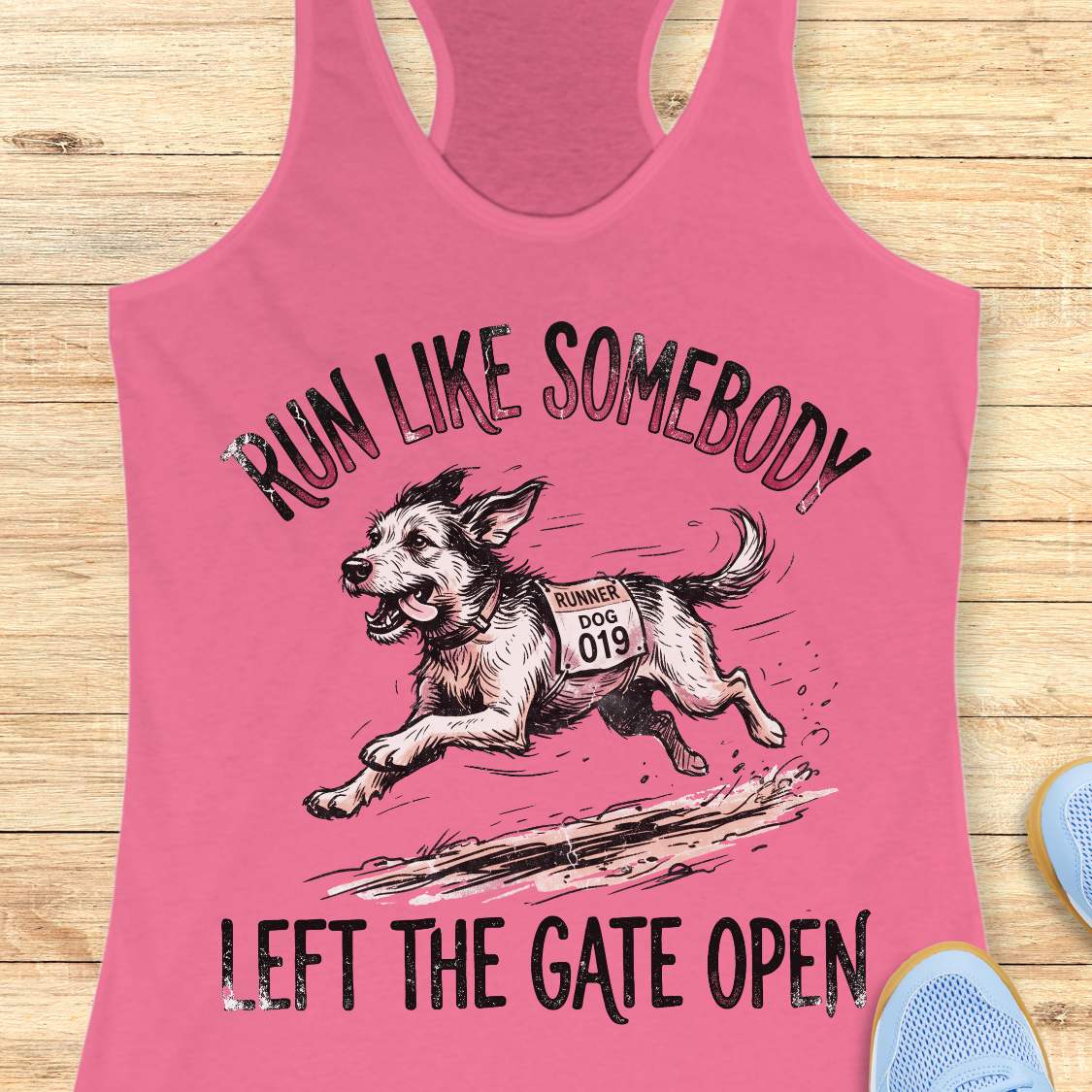 A pink racerback tank top laid flat on a light wood surface, featuring a graphic of a dog wearing a race bib and the text 'RUN LIKE SOMEBODY LEFT THE GATE OPEN.' A pair of blue shoes is partially visible at the lower right edge.