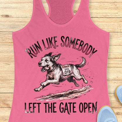 A pink racerback tank top laid flat on a light wood surface, featuring a graphic of a dog wearing a race bib and the text 'RUN LIKE SOMEBODY LEFT THE GATE OPEN.' A pair of blue shoes is partially visible at the lower right edge.