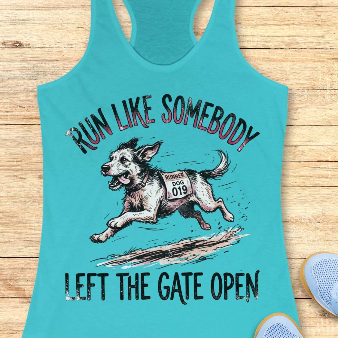 A turquoise racerback tank top laid flat on a light wood surface, featuring a graphic of a running dog with the text 'RUN LIKE SOMEBODY LEFT THE GATE OPEN.' A partial blue shoe is visible at the lower right edge.