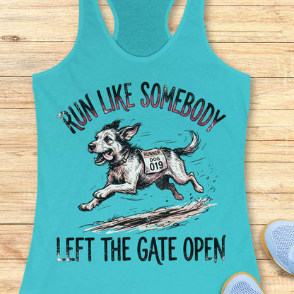 A turquoise racerback tank top laid flat on a light wood surface, featuring a graphic of a running dog with the text 'RUN LIKE SOMEBODY LEFT THE GATE OPEN.' A partial blue shoe is visible at the lower right edge.