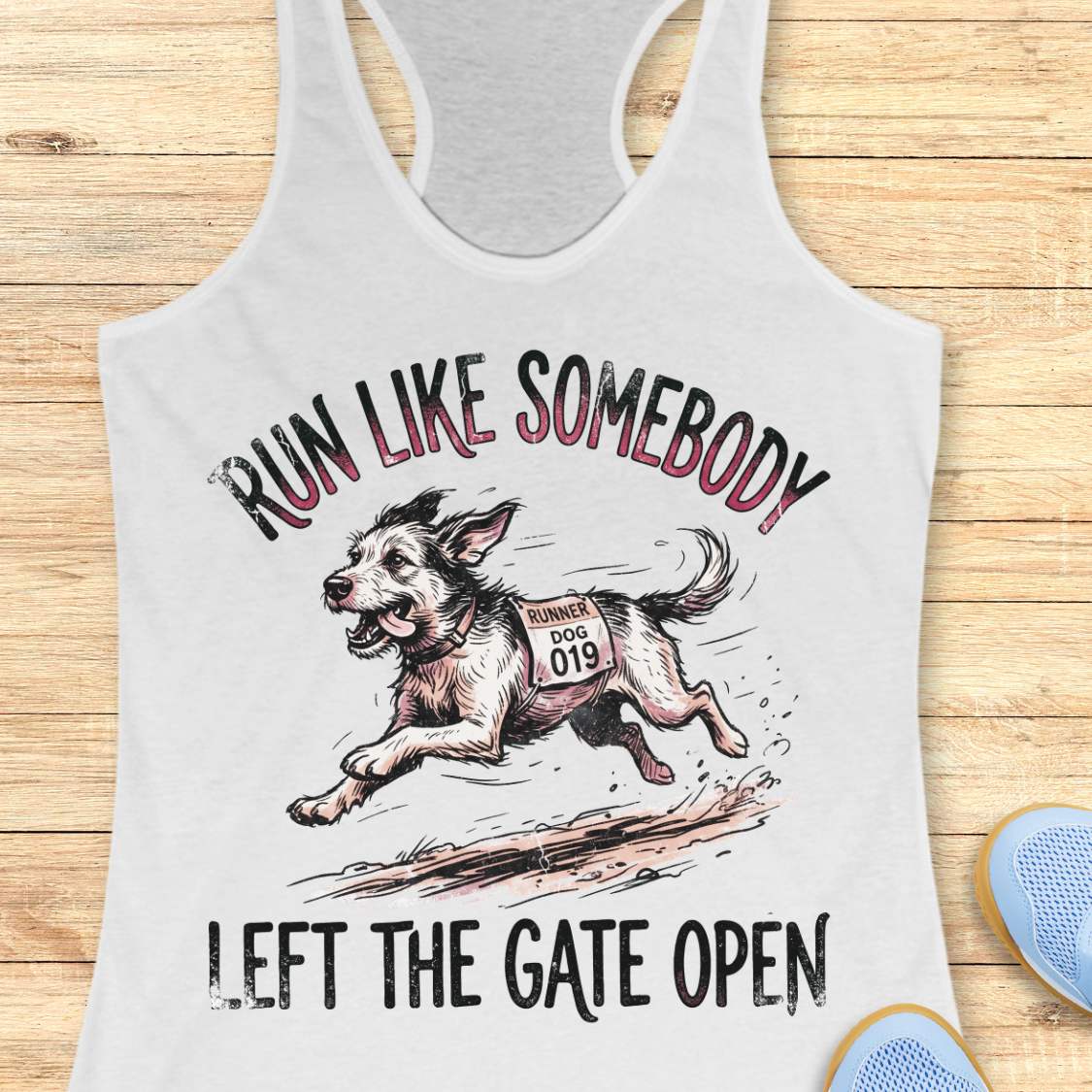 A white racerback tank top laid flat on a light wood background, featuring a graphic of a running dog with the text 'RUN LIKE SOMEBODY LEFT THE GATE OPEN.' A small blue shoe is partially visible at the bottom right edge.