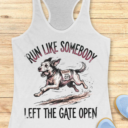A white racerback tank top laid flat on a light wood background, featuring a graphic of a running dog with the text 'RUN LIKE SOMEBODY LEFT THE GATE OPEN.' A small blue shoe is partially visible at the bottom right edge.