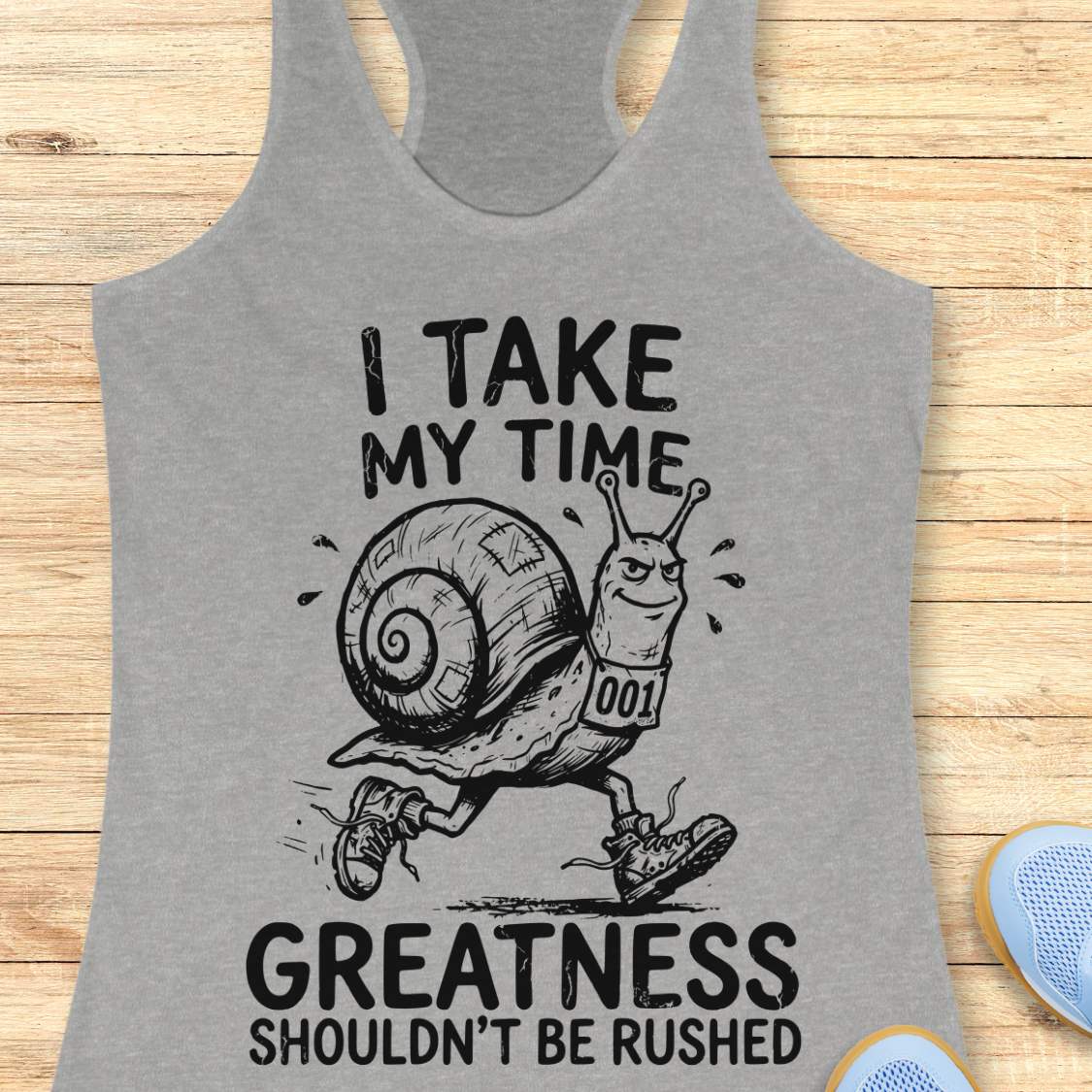 A heather gray racerback tank top laid flat on a light wood background, featuring a black graphic of a snail runner with the text 'I TAKE MY TIME GREATNESS SHOULDN'T BE RUSHED'.