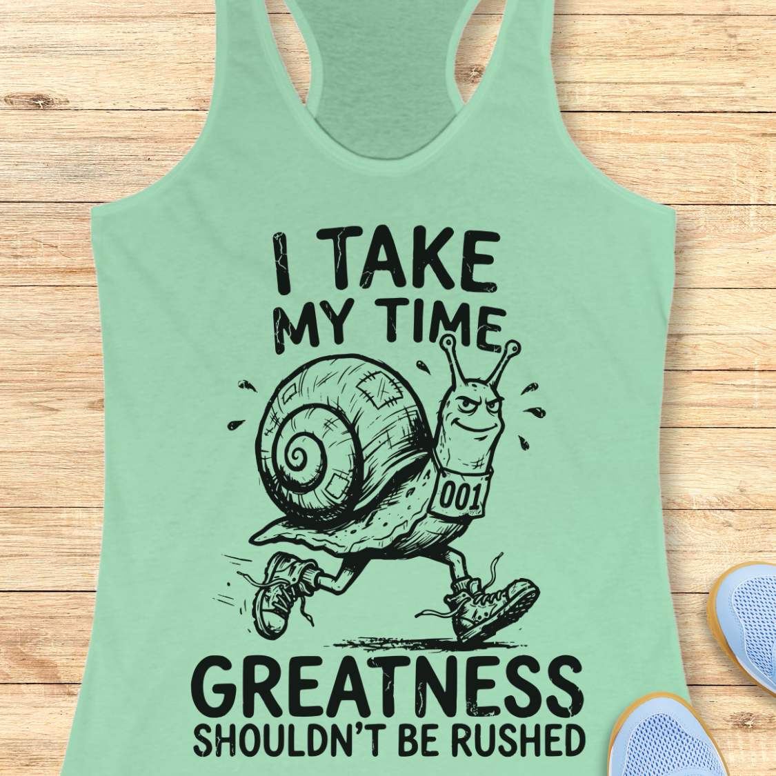 A mint green racerback tank top laid flat on a light wood background, featuring a black graphic of a snail running with the text 'I TAKE MY TIME GREATNESS SHOULDN'T BE RUSHED'.