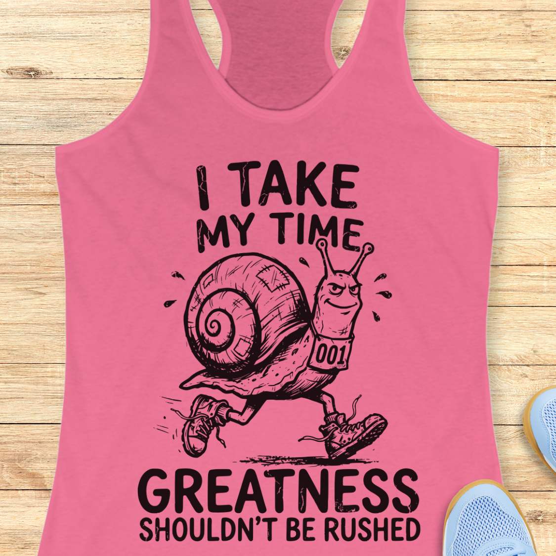 A pink racerback tank top laid flat on a light wood background, featuring a black graphic of a snail runner with the text 'I TAKE MY TIME GREATNESS SHOULDN'T BE RUSHED'.