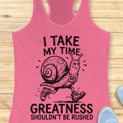 A pink racerback tank top laid flat on a light wood background, featuring a black graphic of a snail runner with the text 'I TAKE MY TIME GREATNESS SHOULDN'T BE RUSHED'.