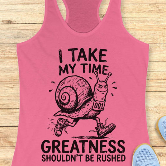 A pink racerback tank top laid flat on a light wood background, featuring a black graphic of a snail runner with the text 'I TAKE MY TIME GREATNESS SHOULDN'T BE RUSHED'.