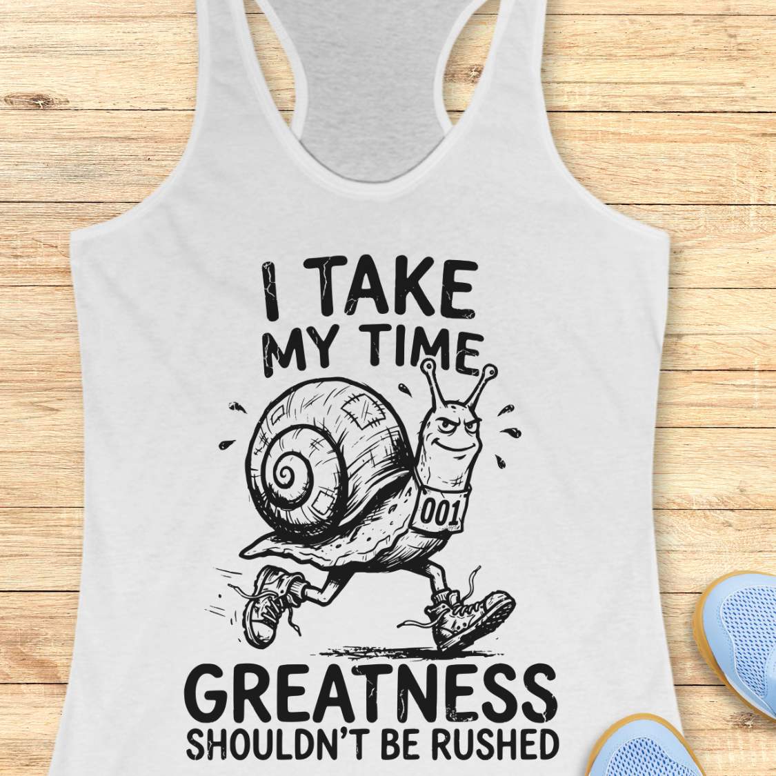 A white racerback tank top laid flat on a light wood background, featuring a black graphic of a snail running with the text 'I TAKE MY TIME GREATNESS SHOULDN'T BE RUSHED'. Partial blue shoes are visible at the bottom right edge.