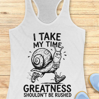 A white racerback tank top laid flat on a light wood background, featuring a black graphic of a snail running with the text 'I TAKE MY TIME GREATNESS SHOULDN'T BE RUSHED'. Partial blue shoes are visible at the bottom right edge.