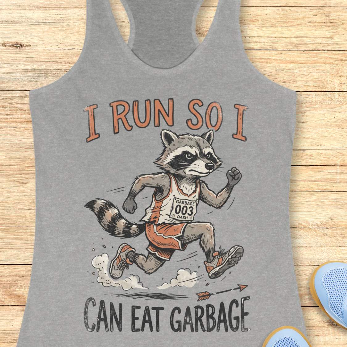 A heather gray racerback tank top laid flat on a light wood background, featuring a graphic of a raccoon runner with the text 'I RUN SO I CAN EAT GARBAGE'.