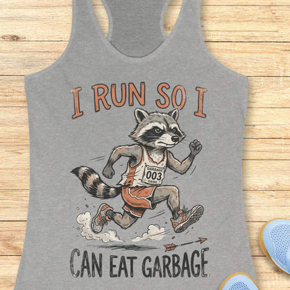A heather gray racerback tank top laid flat on a light wood background, featuring a graphic of a raccoon runner with the text 'I RUN SO I CAN EAT GARBAGE'.