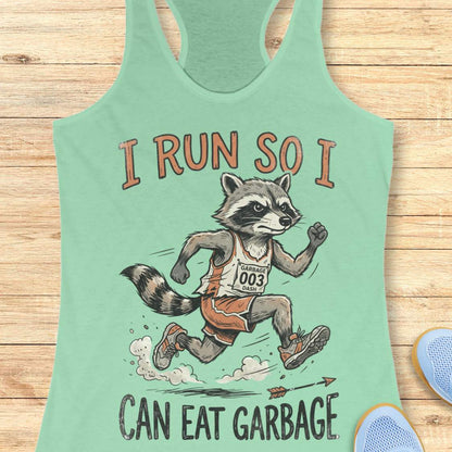 A mint green racerback tank top laid flat on a light wood background, featuring a cartoon raccoon running with the text 'I RUN SO I CAN EAT GARBAGE'. A pair of blue sneakers is partially visible at the bottom right.
