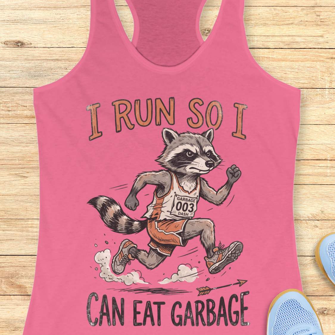 A pink racerback tank top laid flat on a light wood background, featuring a graphic of a raccoon runner with the text 'I RUN SO I CAN EAT GARBAGE'.