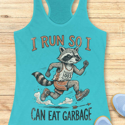 A turquoise racerback tank top laid flat on a light wood surface, featuring a cartoon raccoon running with the text 'I RUN SO I CAN EAT GARBAGE.' A partial blue shoe is visible at the lower right edge.