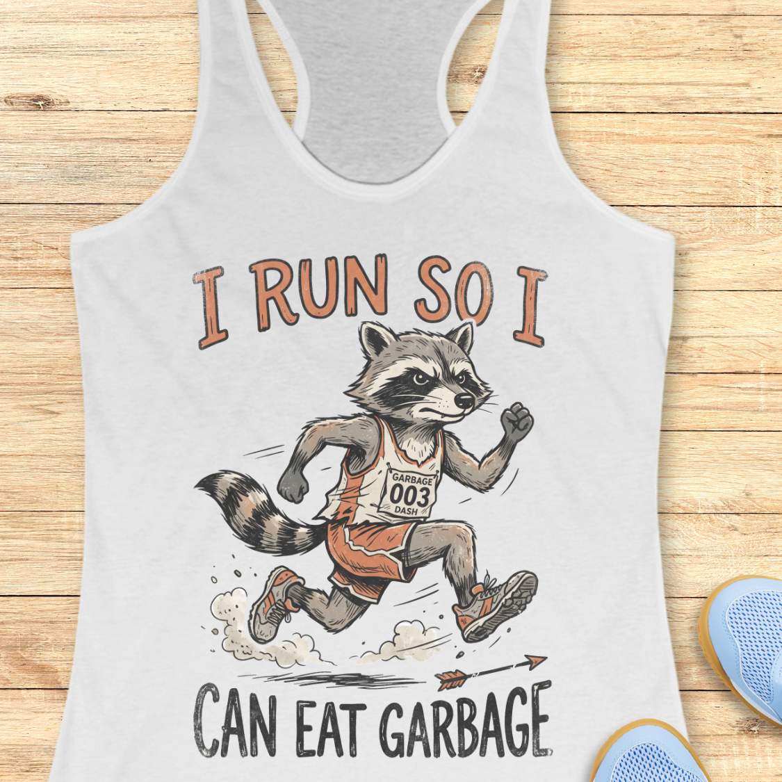 A white racerback tank top laid flat on a light wood background, featuring a printed graphic of a raccoon running in a race with the text 'I RUN SO I CAN EAT GARBAGE'. Part of a blue shoe is visible in the bottom right corner.