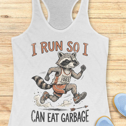A white racerback tank top laid flat on a light wood background, featuring a printed graphic of a raccoon running in a race with the text 'I RUN SO I CAN EAT GARBAGE'. Part of a blue shoe is visible in the bottom right corner.