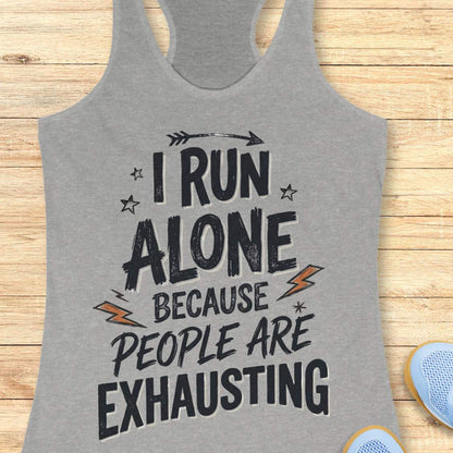 A heather gray racerback tank top with a distressed black graphic text reading 'I RUN ALONE BECAUSE PEOPLE ARE EXHAUSTING' styled on a light wood background with partial blue sneakers visible at the edges.