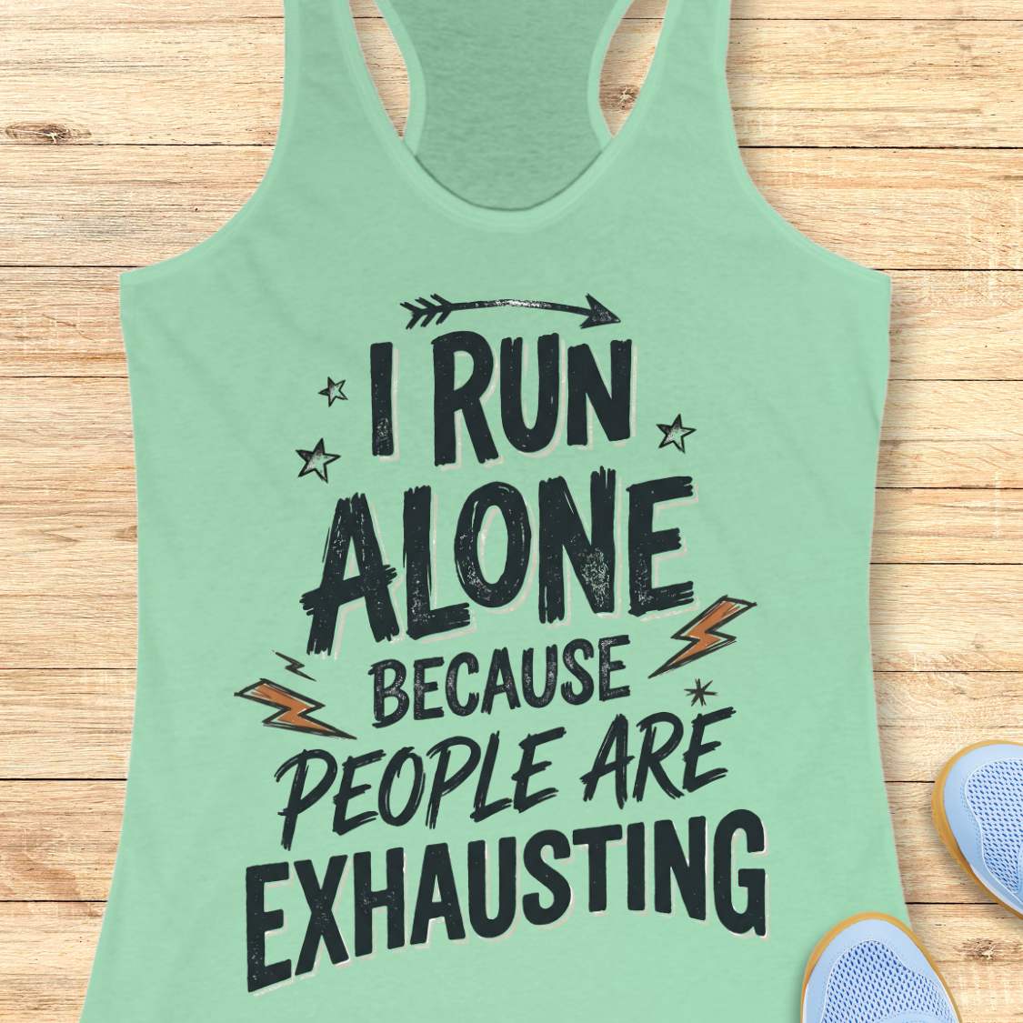 A mint green racerback tank top with a distressed black graphic text that reads 'I RUN ALONE BECAUSE PEOPLE ARE EXHAUSTING' with small orange lightning bolts and black stars, displayed on a light wood background with partial blue sneakers visible at the bottom right.