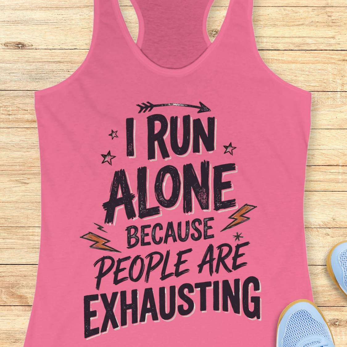 A pink racerback tank top laid flat on a light wood background, featuring a distressed black graphic that reads 'I RUN ALONE BECAUSE PEOPLE ARE EXHAUSTING' with small star and lightning accents. Partial blue shoes are visible at the bottom right as styling props.