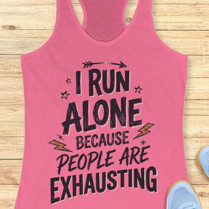 A pink racerback tank top laid flat on a light wood background, featuring a distressed black graphic that reads 'I RUN ALONE BECAUSE PEOPLE ARE EXHAUSTING' with small star and lightning accents. Partial blue shoes are visible at the bottom right as styling props.