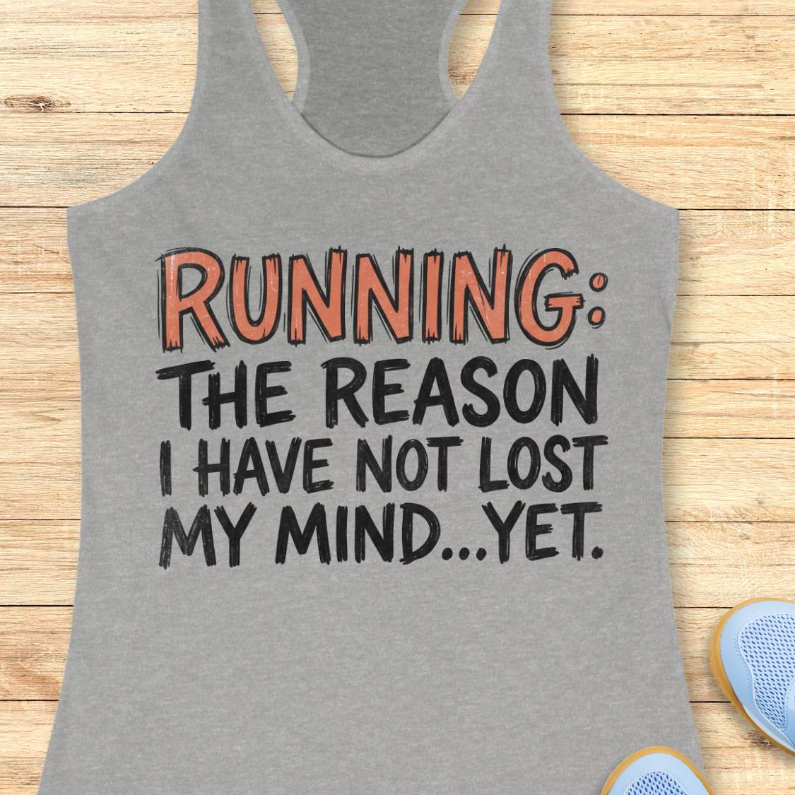 A heather gray racerback tank top laid flat on a light wood background, featuring the printed slogan 'RUNNING: THE REASON I HAVE NOT LOST MY MIND.