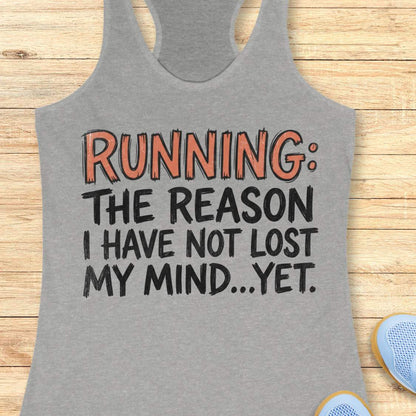 A heather gray racerback tank top laid flat on a light wood background, featuring the printed slogan 'RUNNING: THE REASON I HAVE NOT LOST MY MIND.