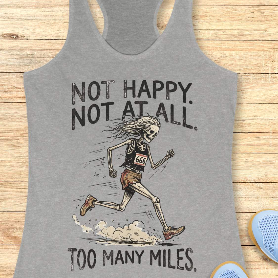 A heather gray racerback tank top laid flat on a light wood surface, featuring a distressed black graphic of a running skeleton with the text 'NOT HAPPY. NOT AT ALL.