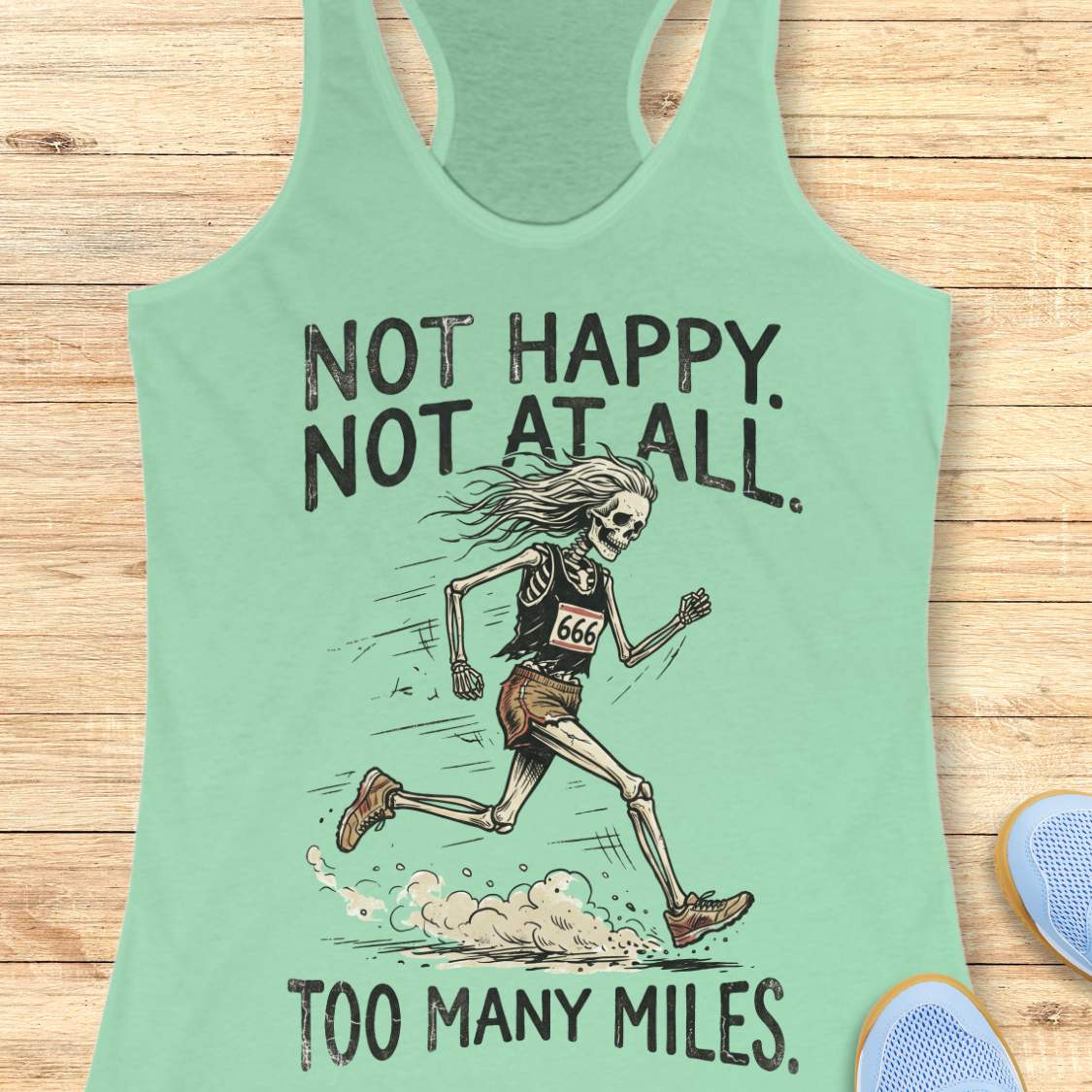 A mint green racerback tank top laid flat on a light wood background, featuring a graphic of a running skeleton with the text 'NOT HAPPY. NOT AT ALL.