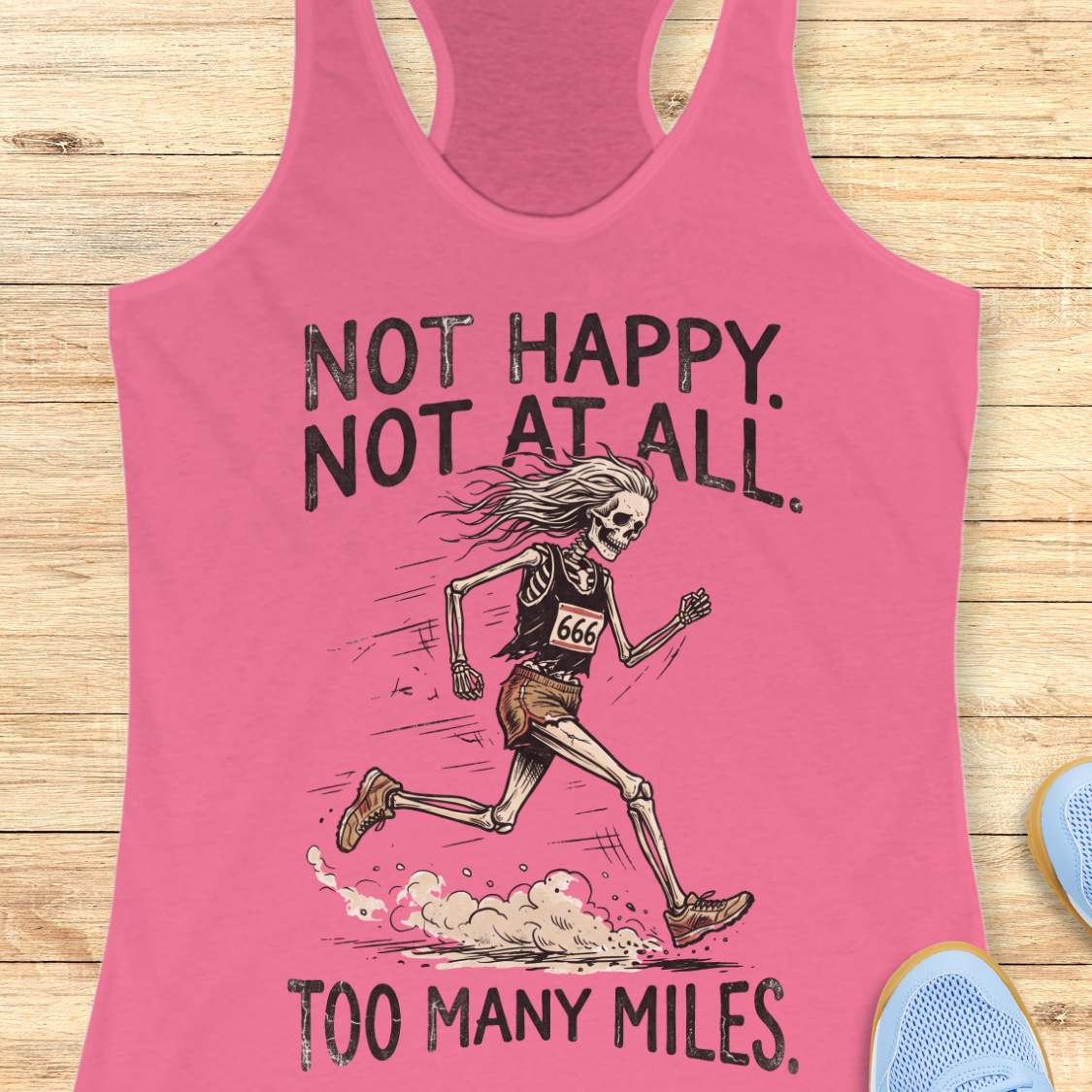 A pink racerback tank top laid flat on a light wood surface, featuring a distressed black graphic of a running skeleton with the text 'NOT HAPPY. NOT AT ALL.