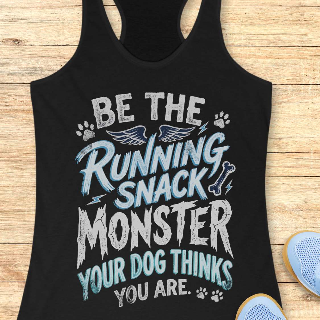 A black racerback tank top laid flat on a light wood background, featuring a humorous graphic that reads 'BE THE RUNNING SNACK MONSTER YOUR DOG THINKS YOU ARE.' in white and light blue lettering with paw prints, wings, and lightning accents.