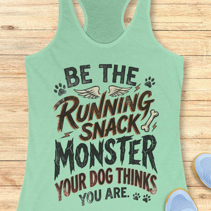A mint green racerback tank top laid flat on a light wood background, featuring a humorous graphic print that reads 'BE THE RUNNING SNACK MONSTER YOUR DOG THINKS YOU ARE.' with dog-themed illustrations including paw prints, wings, a bone, and lightning bolts.
