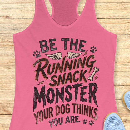 A pink racerback tank top with a humorous graphic that reads 'BE THE RUNNING SNACK MONSTER YOUR DOG THINKS YOU ARE.' in distressed black lettering, styled flat on a light wood background with a shoe partially visible in the corner.